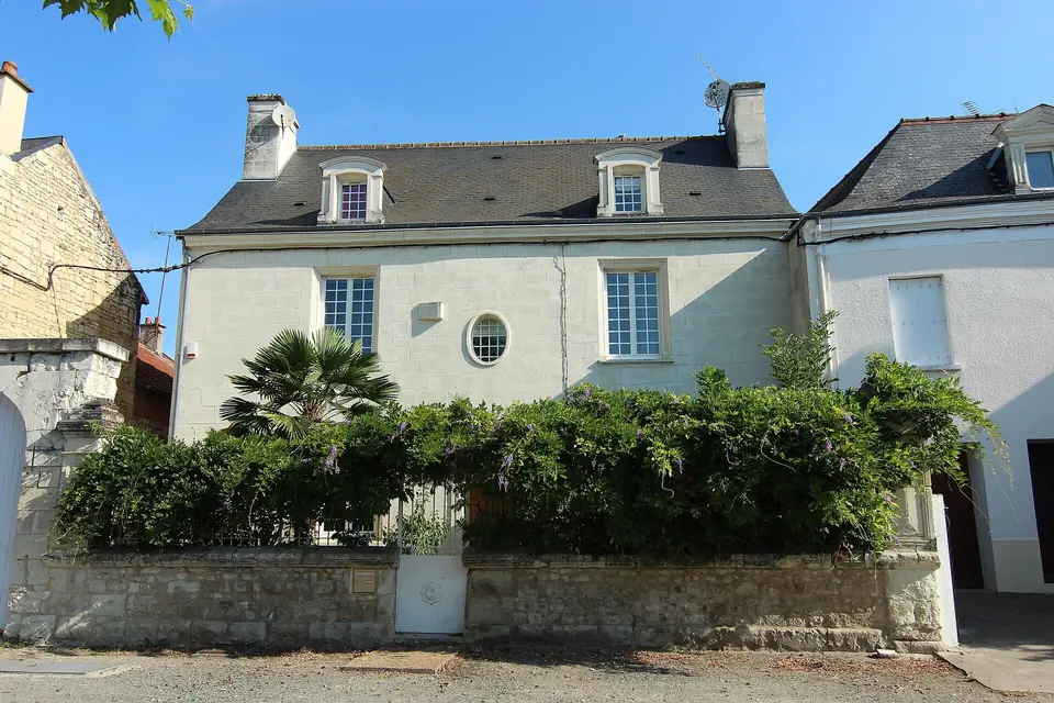 Maison bourgeoise blanche à Thuré avec fenêtres à petits carreaux, lucarnes en toiture et oculus, derrière un mur couvert de végétation.