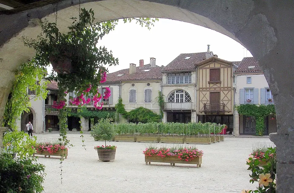 Place Royale de Labastide-d'Armagnac vue depuis les arcades, avec maisons à colombages et façades claires ornées de jardinières fleuries.